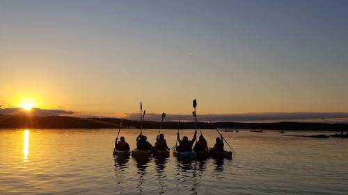 Silhouetted kayakers paddle in the water at sunset, with a vibrant sky and calm reflections on the surface.