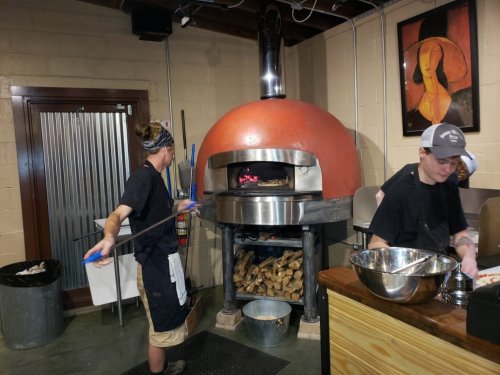 Two chefs working in a restaurant kitchen, one placing a pizza in a large wood-fired oven.