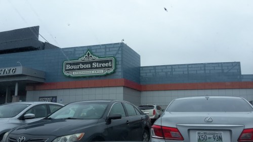 Exterior of Bourbon Street Steakhouse & Grill, with a cloudy sky and parked cars in the foreground.