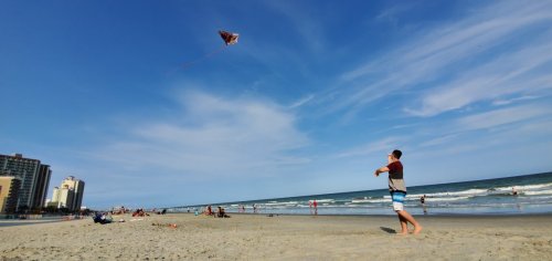 A child flying a kite on a beach with waves and people in the background under a clear blue sky.