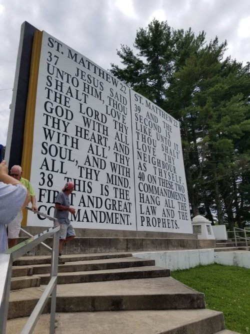 A large sign displays biblical verses from St. Matthew, with people sitting nearby on steps under a cloudy sky.