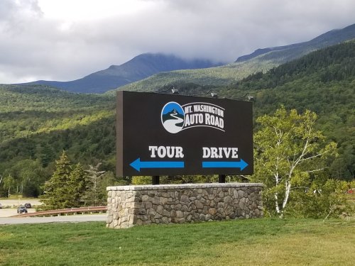 Sign for Mt. Washington Auto Road with arrows pointing left for "Tour" and right for "Drive," set against a mountain backdrop.