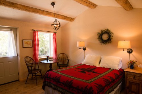 Cozy bedroom with a red patterned blanket, wooden beams, a round table, and warm lighting from lamps.