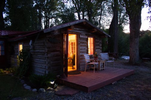 A cozy log cabin at dusk, with warm light glowing from the windows and two chairs on the porch.