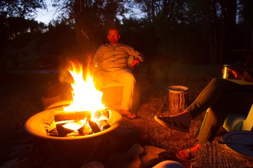 A person sits by a campfire at dusk, enjoying snacks, with a dog nearby and another person relaxing in the background.