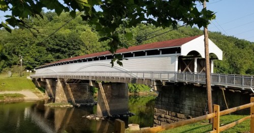 A white covered bridge spans a calm river, surrounded by green trees and a clear blue sky.