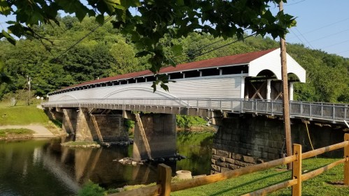 A white covered bridge spans a calm river, surrounded by green trees and a wooden fence in the foreground.