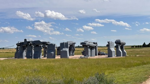 A sculpture featuring stacked gray cars on stone-like structures, set against a blue sky and grassy landscape.