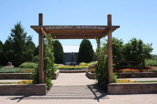 Entrance to Sensory Gardens featuring a wooden archway, colorful flower beds, and lush greenery under a clear blue sky.
