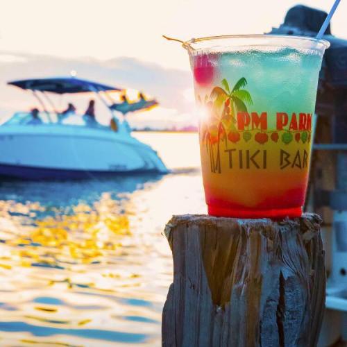 A colorful drink with a cherry sits on a post by the water, with a boat in the background and a sunset glow.