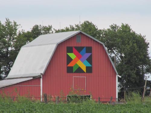 A red barn with a colorful geometric quilt pattern on its side, surrounded by green trees and fields.