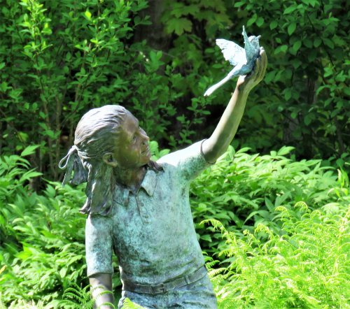 A bronze statue of a girl holding a bird in her hand, surrounded by lush green foliage.