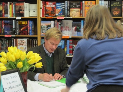 An author signs books at a table, surrounded by shelves of books and a bouquet of yellow tulips in the foreground.