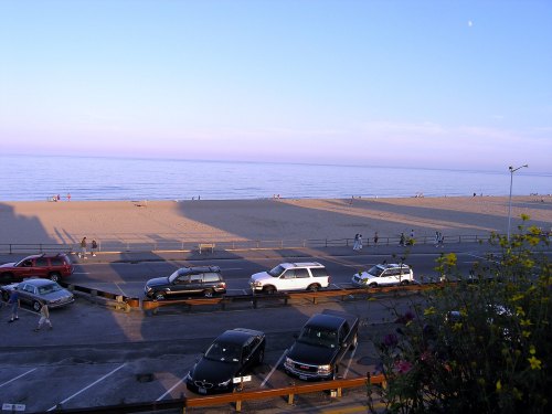 A beach scene at sunset with cars parked along the road and people walking on the sand.