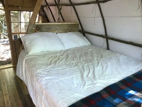 Cozy interior of a rustic cabin featuring a wooden bed with white bedding and a plaid blanket. Natural light filters in.