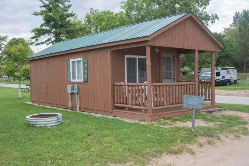 A small brown cabin with a green metal roof, porch, and grassy area, surrounded by trees and a campsite.