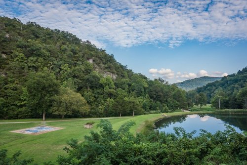 A serene landscape featuring a pond, lush greenery, and rolling hills under a partly cloudy sky.