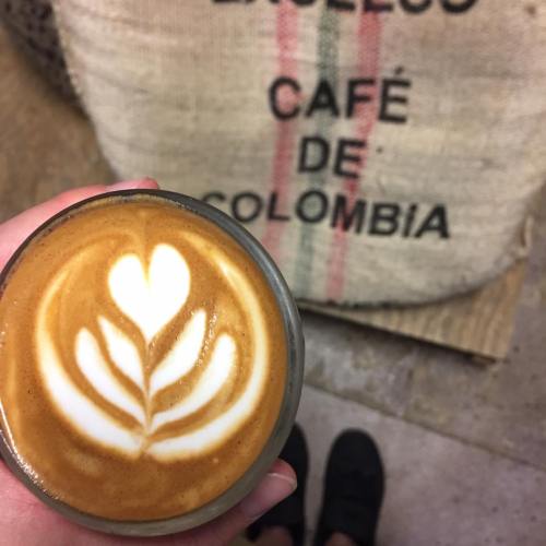 A hand holding a latte with heart-shaped foam art in front of a burlap sack labeled "Café de Colombia."