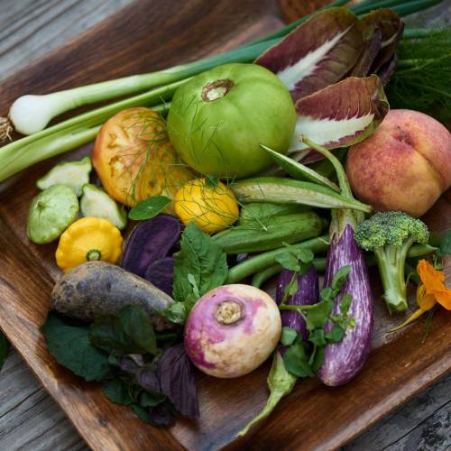 A wooden tray filled with a variety of fresh vegetables and fruits, including greens, squash, and a peach.
