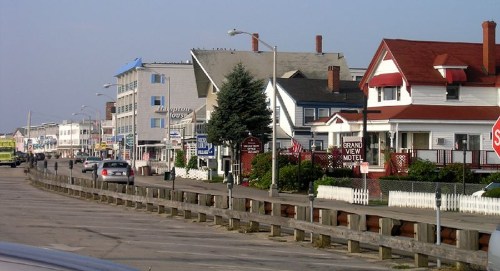 A coastal street lined with various buildings, including motels and shops, with cars parked along the road.