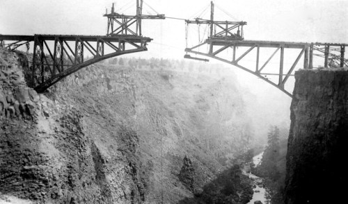 Black and white photo of an incomplete bridge spanning a deep canyon, with construction supports visible.