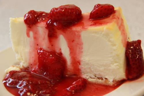 A slice of cheesecake topped with a glossy strawberry sauce and fresh strawberries on a white plate.