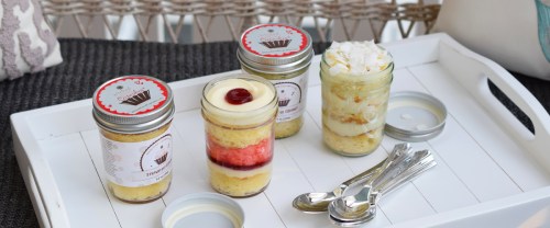 Four layered desserts in jars on a white tray, featuring colorful layers and decorative lids, with spoons beside them.