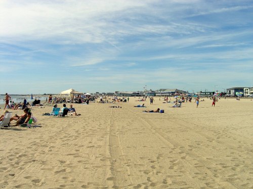 A sunny beach scene with people lounging on sand, umbrellas, and a distant boardwalk under a clear blue sky.