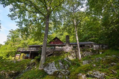 A rustic cabin nestled among trees, featuring a stone chimney and a wooden deck, surrounded by lush greenery.