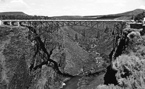 A historic black-and-white photo of a large metal bridge spanning a deep canyon with a river below.
