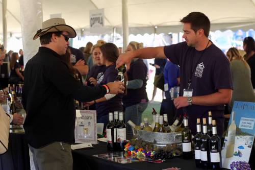 A man in a hat receives a wine pour from a vendor at a wine tasting event under a tent.