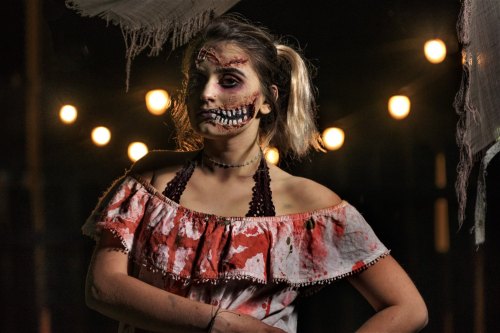 A girl with a spooky makeup design, featuring a skull face and blood-stained clothing, stands under dim lights.