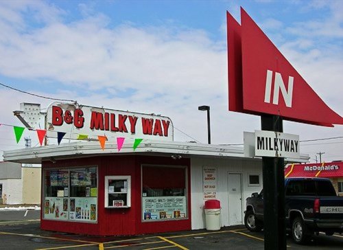 A small, colorful ice cream shop with a red roof and sign, surrounded by a parking lot and festive banners.