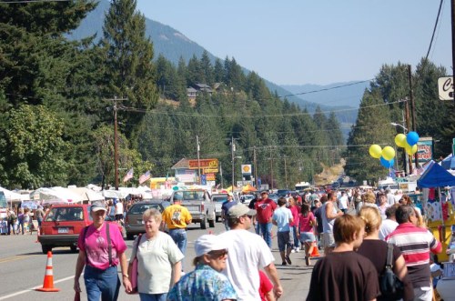A busy street fair with people walking, food stalls, and colorful balloons, set against a backdrop of trees and mountains.