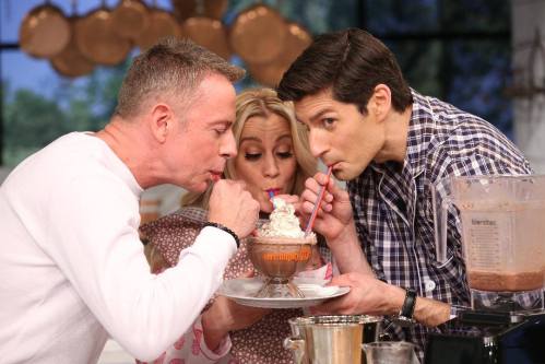 Three people share a dessert, sipping from straws in a bowl, with a blender and kitchen backdrop.