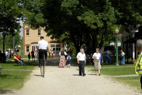 A tall stilt walker in a park surrounded by people, trees, and historic buildings on a sunny day.