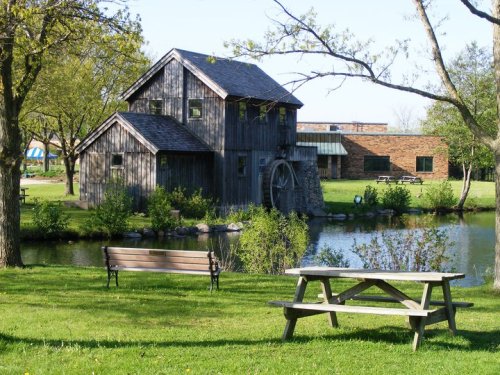A rustic wooden building with a water wheel beside a pond, surrounded by greenery and picnic tables.