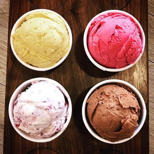 Four ice cream scoops in cups: yellow, pink, white with swirls, and brown, arranged on a wooden tray.