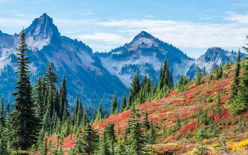 A scenic view of mountains with rugged peaks, surrounded by vibrant red foliage and evergreen trees under a blue sky.