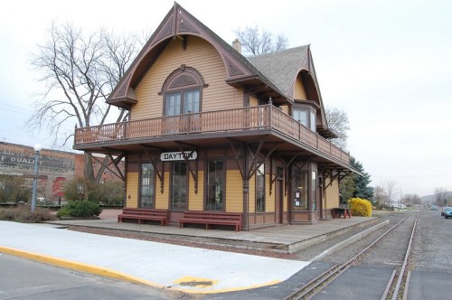 Historic train station with a wooden exterior, featuring a balcony and benches, set along a railway track.