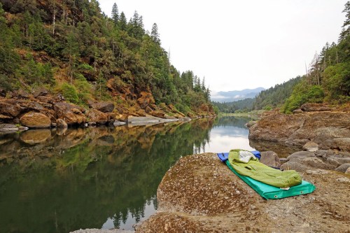 A serene river scene with lush green trees, rocky banks, and a sleeping bag laid out on a large rock by the water.