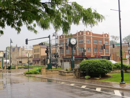 A rainy street scene featuring a clock tower, brick buildings, and traffic lights in a small town.
