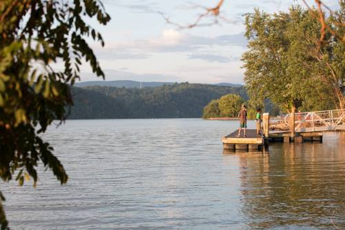 Two people stand on a wooden dock by a calm lake, surrounded by trees and mountains in the background.