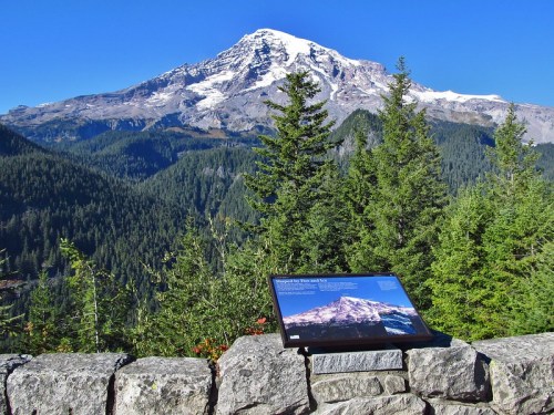 A scenic view of a snow-capped mountain surrounded by lush green forests, with an informational sign in the foreground.