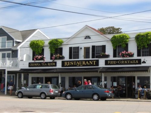 Exterior of the Olympia Tea Room restaurant, featuring a black sign, flower boxes, and parked cars in front.