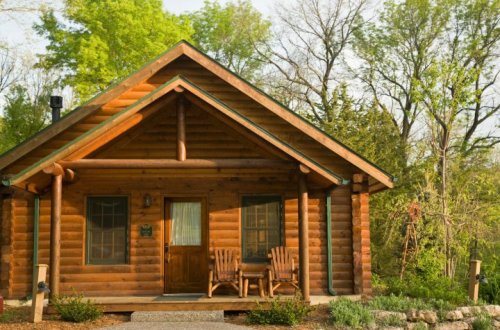A cozy log cabin surrounded by greenery, featuring a front porch with two wooden chairs.