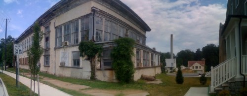 Abandoned building with large windows, overgrown vegetation, and a cloudy sky in the background.