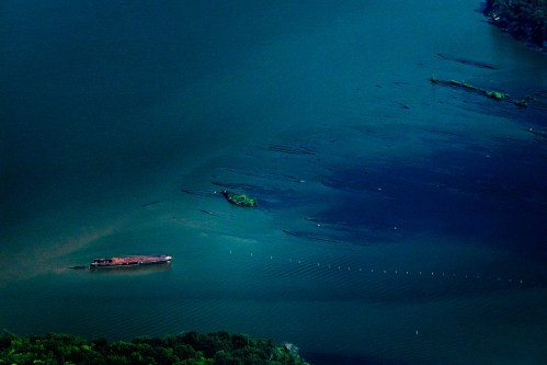 Aerial view of a ship on dark blue water, with submerged vegetation and a shoreline in the background.