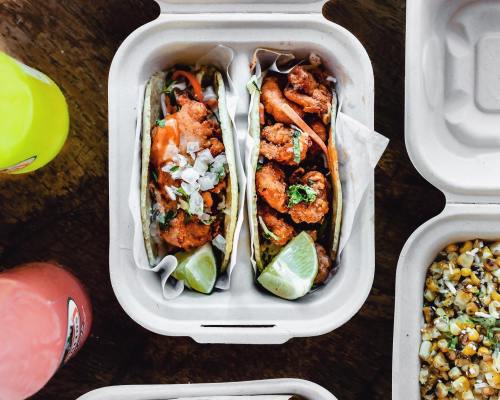 Two tacos filled with crispy fried shrimp, topped with onions and cilantro, served with lime wedges in a takeout container.