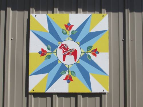 Colorful barn quilt featuring a red horse in the center, surrounded by blue and yellow rays and red roses.